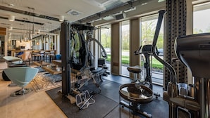 A modern gym with various exercise equipment, a dining area, and large windows offering views of the outdoors. - MASEVEN München Messe Trudering (Munich)