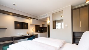 A modern hotel room with a large bed, a desk with a chair, a flat-screen TV, and a wardrobe. - MASEVEN München Messe Trudering (Munich)