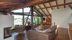A spacious living area with a high ceiling, wooden beams, a large window, and a fireplace.