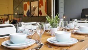A dining table set with white bowls, plates, and cutlery, with a potted plant and framed artwork on the wall behind.