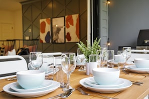 Una mesa de comedor dispuesta con cuencos, platos y cubiertos blancos, con una planta en maceta y una obra de arte enmarcada en la pared de fondo.