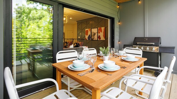 A dining area with a wooden table set for four, white chairs, and a view of the outdoors through a glass door.