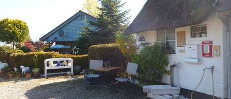 A rustic building with a thatched roof, a small outdoor seating area with a bench and table, and a gravel ground.