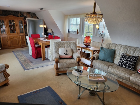Una sala de estar acogedora con una mesa de centro de vidrio, un sofá con cojines estampados, un aparador de madera y un comedor con una silla roja.