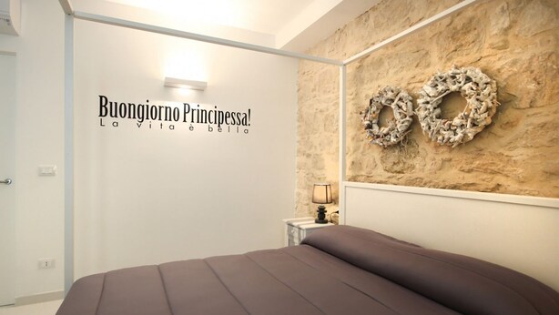 A bedroom with a bed, a nightstand, a wall-mounted lamp, and two wreaths on the wall. - Just 100 meters from one of the most beautiful and renowned beaches of the Ragusa’s coast (Marina di Ragusa)