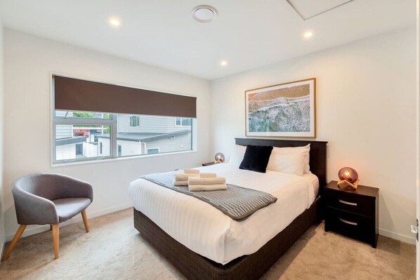 Un dormitorio moderno con una cama grande, una silla, una mesita de noche y una ventana con vistas a edificios.