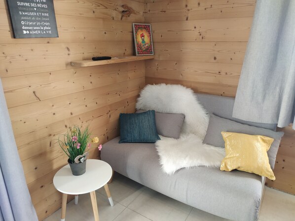 Una habitación acogedora con pared de madera, un sofá gris con cojines blancos y amarillos, una mesita pequeña con planta en maceta y un cuadro enmarcado sobre el sofá.