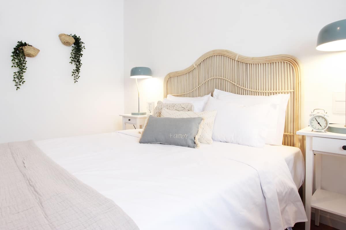 Una cama bien hecha con ropa de cama blanca y almohada gris, una mesita de noche con lámpara y un reloj de pared.