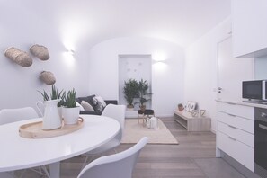 Un espacio moderno y minimalista para sala y comedor con una mesa redonda, sillas blancas, un sofá y una cocina con televisor y armarios.