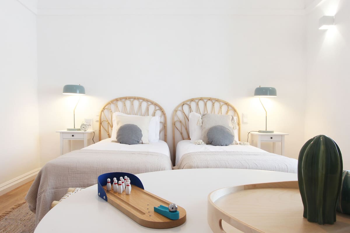 Un dormitorio con dos camas, mesitas de noche, lámparas, una bandeja de madera con bolos y un jarrón verde grande.