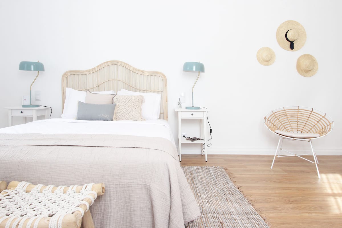 Un dormitorio ordenado con cama, mesitas de noche, una silla de mimbre y una pared con tres sombreros.