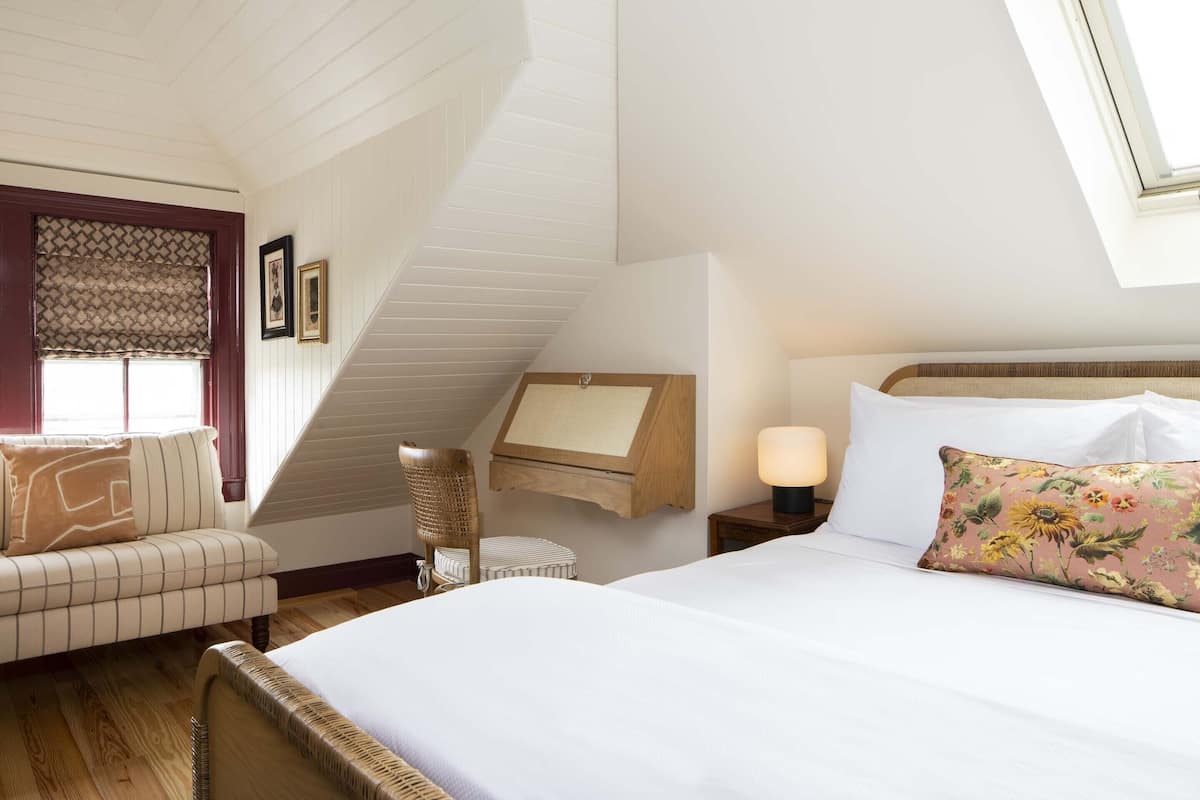 Une chambre avec un plafond en pente, un lit avec du linge de lit blanc, un coussin à motifs floraux, une table de chevet avec une lampe, une chaise et une fenêtre avec des rideaux à motifs.
