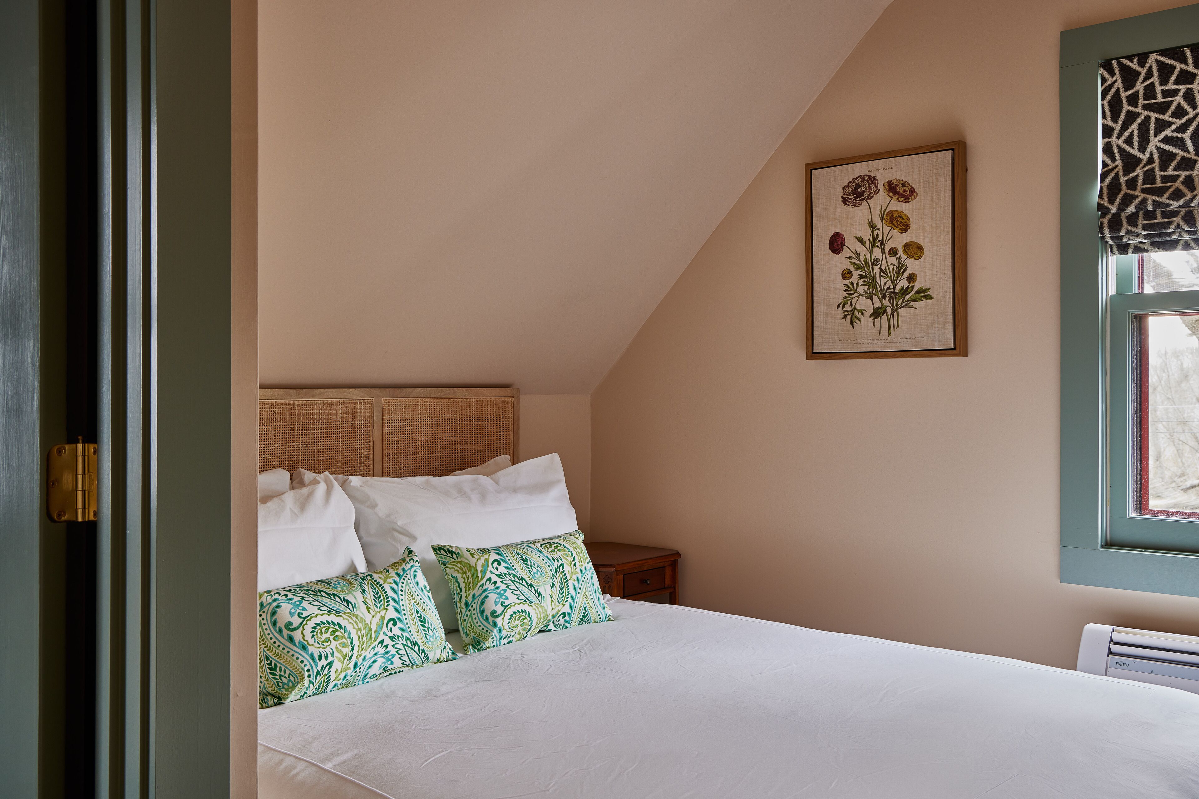 A bedroom with a bed, pillows, a nightstand, a framed floral artwork, and a window with a patterned blind.