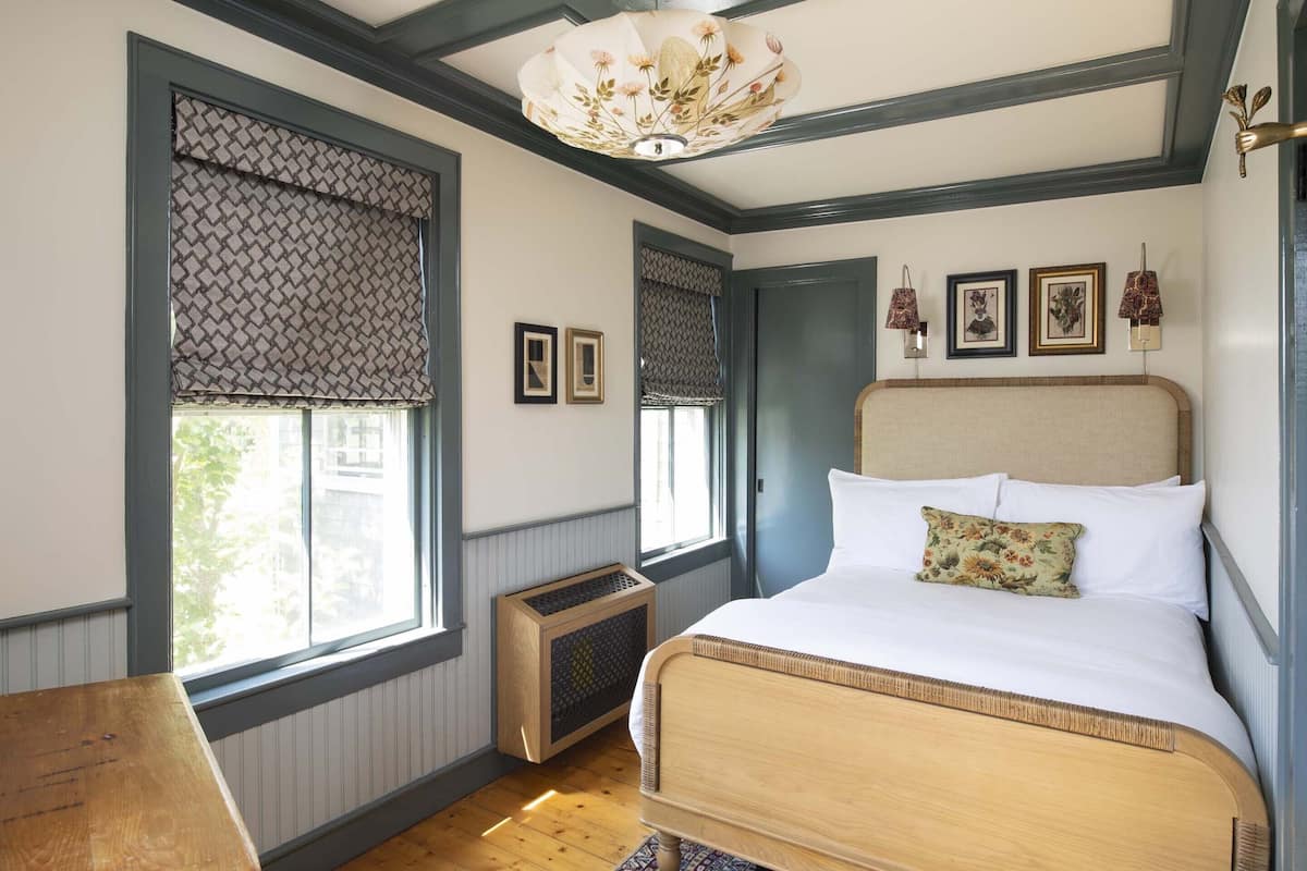 Une chambre avec un grand lit, deux fenêtres équipées de stores à motifs, un radiateur et un luminaire au plafond.