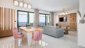 Una sala de estar moderna con sofá, mesa de comedor, sillas, televisor y vista al mar.