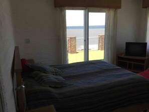 A bedroom with a bed, a television on a wooden stand, and a view of the sea through a sliding door.