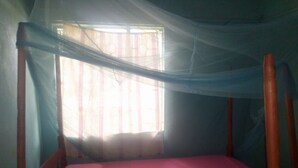 Cama de solteiro com colchão rosa e cabeceira de madeira em quarto com janela coberta por uma cortina translúcida.