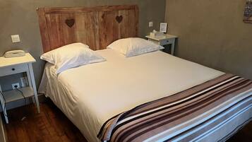 Una cama bien hecha con sábanas blancas y una manta a rayas, un cabecero de madera con formas de corazón, mesitas de noche con lámparas y un teléfono sobre la mesa de la izquierda.