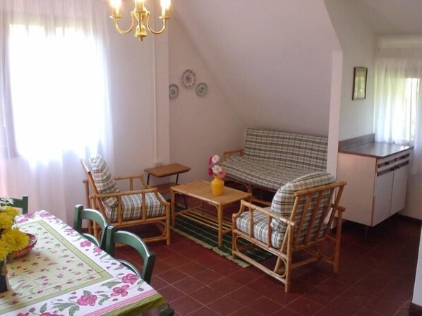 Un salon confortable avec un canapé, deux fauteuils, une petite table et une table à manger avec des chaises. Il y a également un coin cuisine avec un évier et une fenêtre avec des rideaux.