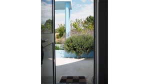 A view through a glass door of a building with a blue structure and a small garden area with bushes and a concrete path.