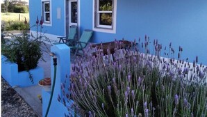 A blue house with three windows, a covered patio with two chairs, and a garden with lavender plants.