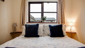 A neatly made bed with a floral patterned bedspread, two dark blue pillows, and a window with curtains. - Hop Cottage (Ashford)