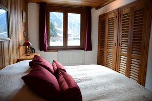 Quarto com cama, janela com cortinas, radiador e persianas de madeira.