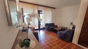 Ein Wohnzimmer mit Sofa, Couchtisch und Esstisch. Ein Fenster mit Vorhängen und eine Tür sind ebenfalls vorhanden.