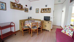 Una habitación pequeña y bien iluminada con una mesa de comedor de madera, dos sillas, un televisor sobre un mueble de madera y un estante metálico rojo.