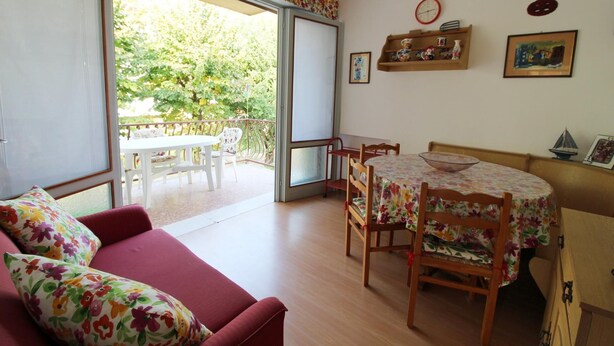 Un salón acogedor con un sofá rojo, una mesa de comedor con manteles florales y un balcón con mesa y sillas blancas.