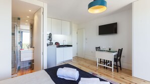Una habitación de hotel moderna con una cama, un pequeño espacio kitchenette, una zona de comedor y un televisor fijado en la pared.