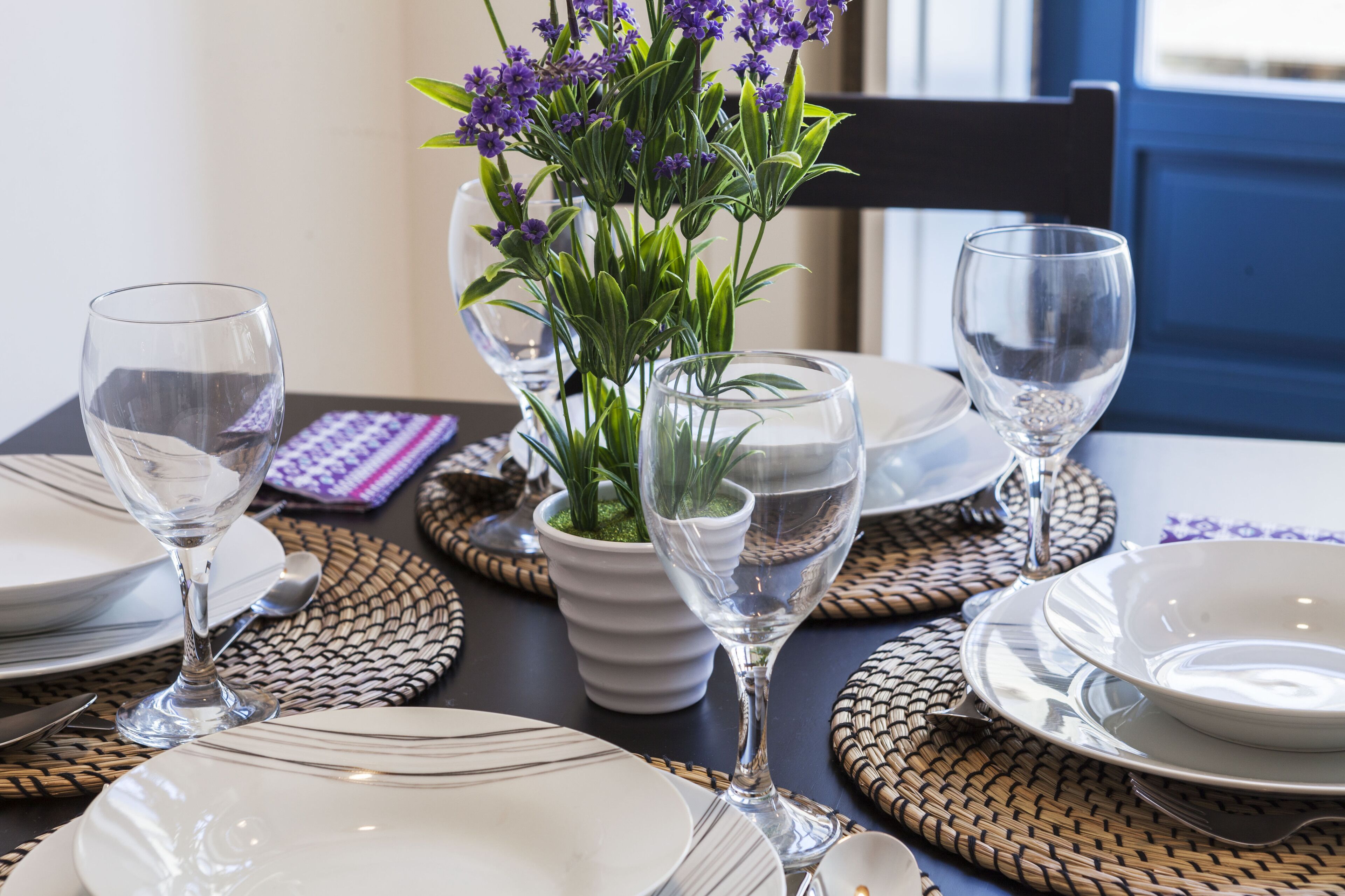 Una mesa de comedor dispuesta con platos blancos, copas de vino transparentes y una planta en maceta con flores moradas.