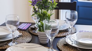 A dining table set with white plates, clear wine glasses, and a potted plant with purple flowers.