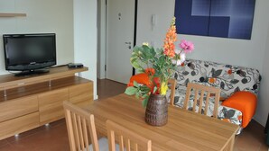 Een eethoek met een houten tafel, stoelen, een vaas met bloemen, een televisie op een kast en een bank met gekleurde kussens.