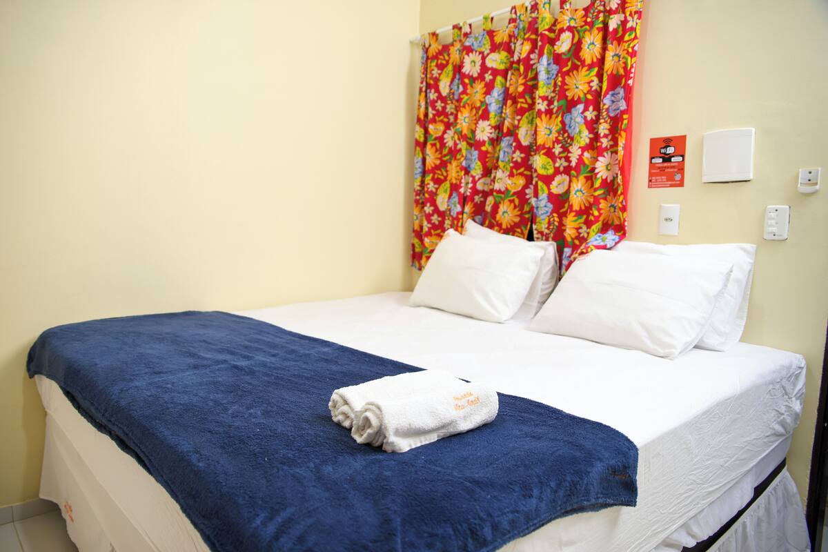 Habitación de hotel con cama, almohadas blancas, manta azul y cortina con estampado floral.