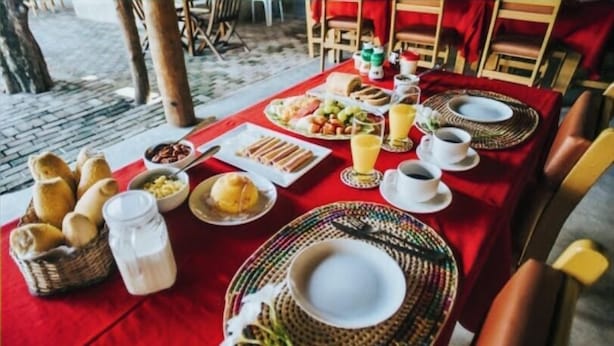 A breakfast setting with a red tablecloth, plates, bread, and beverages. - Pousada Meu Xodó (São Miguel dos Milagres)