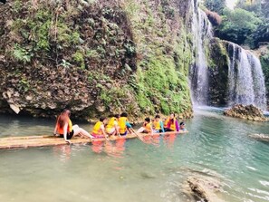 Un grup de persones assegudes sobre una llarga plataforma de fusta en un riu, envoltat de vegetació exuberant i una cascada.