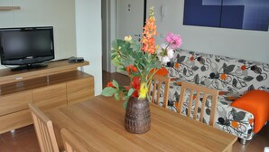 A modern living room with a wooden dining table, a television on a wooden stand, a sofa with a floral pattern, and a vase with flowers.