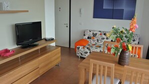 Un salon avec un téléviseur posé sur un meuble en bois, un canapé à motifs floraux, une table à manger avec des chaises et un vase rempli de fleurs.