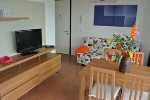 A living room with a TV on a wooden stand, a sofa with a floral pattern, a dining table with chairs, and a vase with flowers.