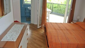 Une chambre avec une commode en bois, un lit recouvert d’une couverture orange, une salle de bain avec un lavabo et un miroir, et un balcon donnant sur un paysage verdoyant.