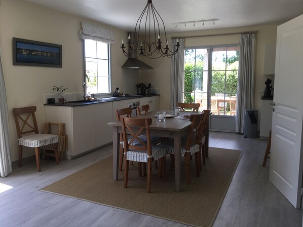 A dining area with a wooden table and chairs, a kitchen with a window, and a view of greenery outside.