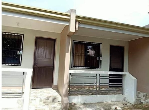 A building with a brown door, two windows with metal bars, and a white railing on a stone-paved area.