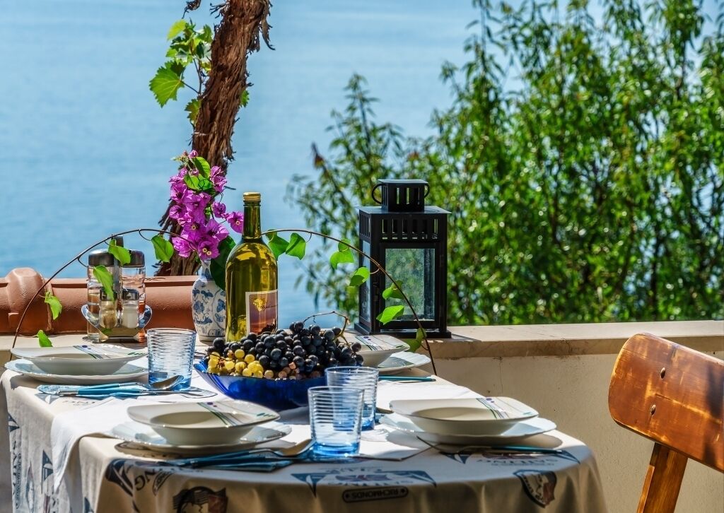 Mesa preparada para comer con platos, vasos y una botella de aceite de oliva, con vista a un cuerpo de agua.
