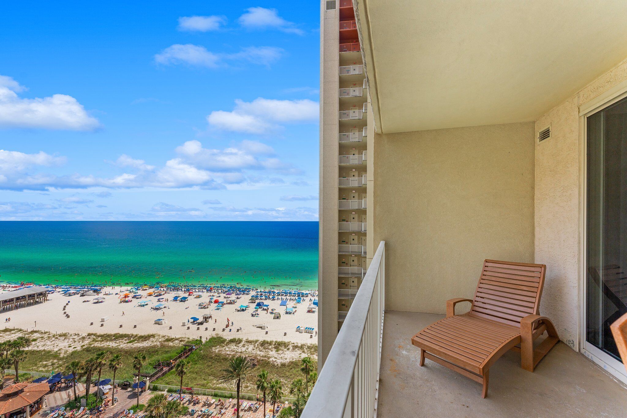 A balcony with a wooden chair, overlooking a beach with umbrellas and people.