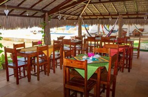 Een terras met houten tafels en stoelen, een rieten dak, en uitzicht op een zwembad en groen.