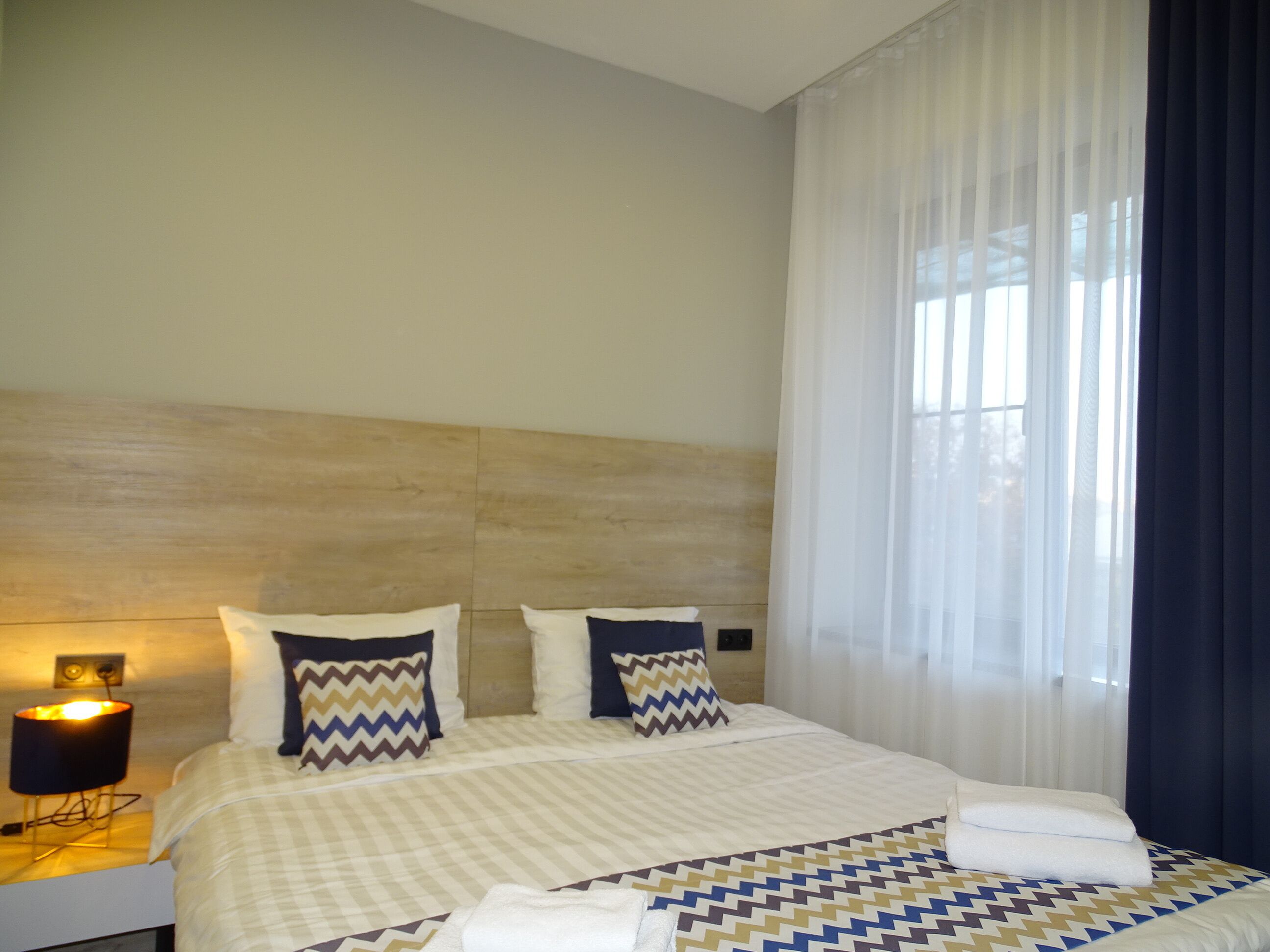 A hotel room with a large bed, two pillows, a nightstand with a lamp, and sheer curtains.
