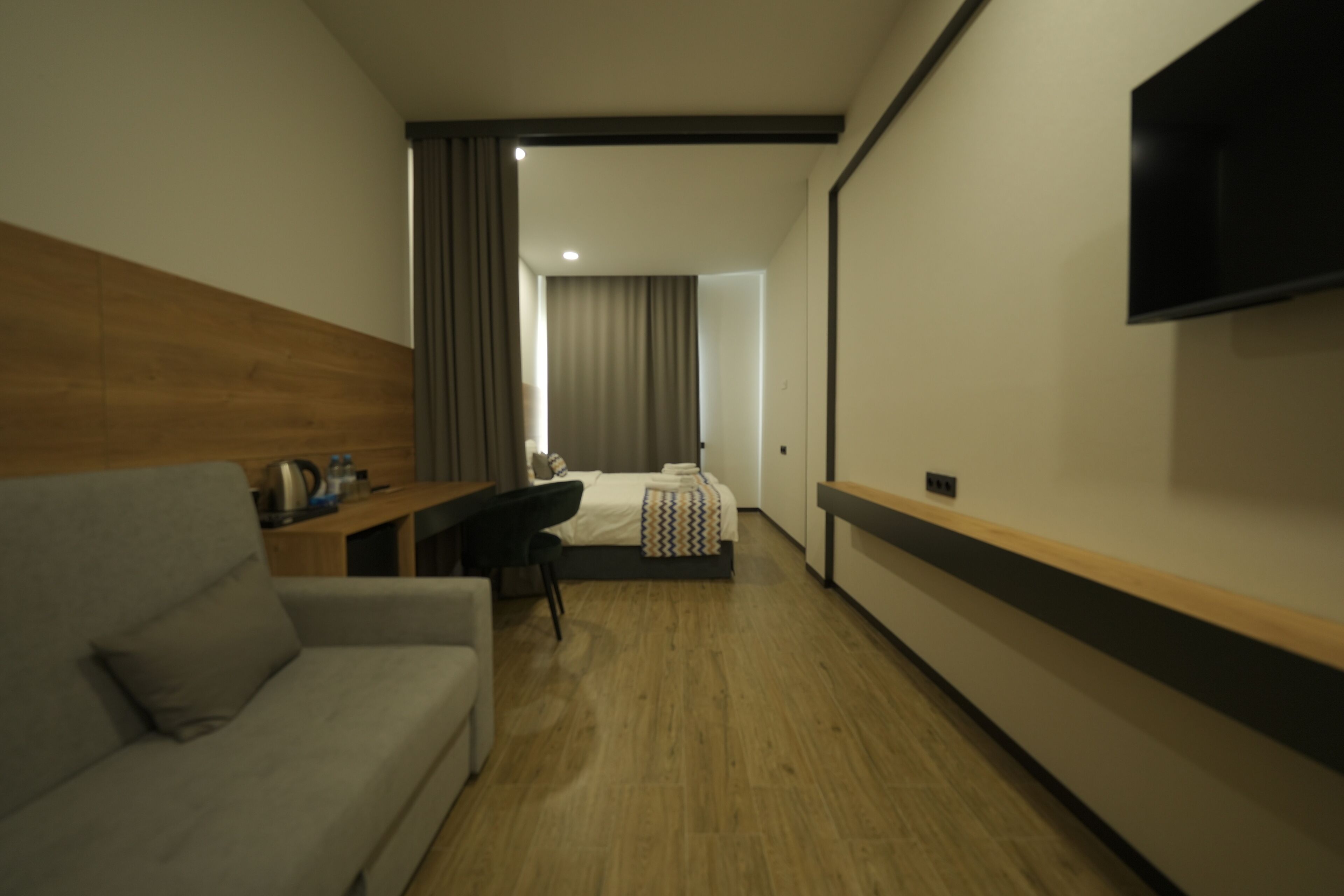 Habitación de hotel con piso de madera, un sofá gris, un escritorio con una silla, una cama con colcha estampada y un televisor de pantalla plana montado en la pared.