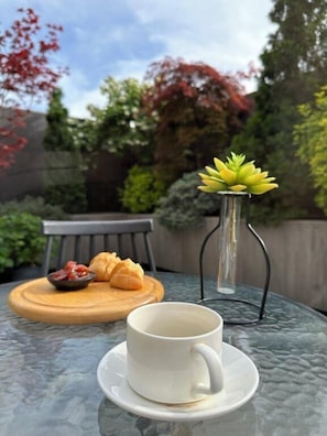 Lasinen pöytä, jonka päällä on valkoinen kuppi ja aluslautanen, puinen tarjoilulauta, jossa on croissantteja ja hilloa, sekä läpinäkyvä maljakko, jossa on mehikasveja.