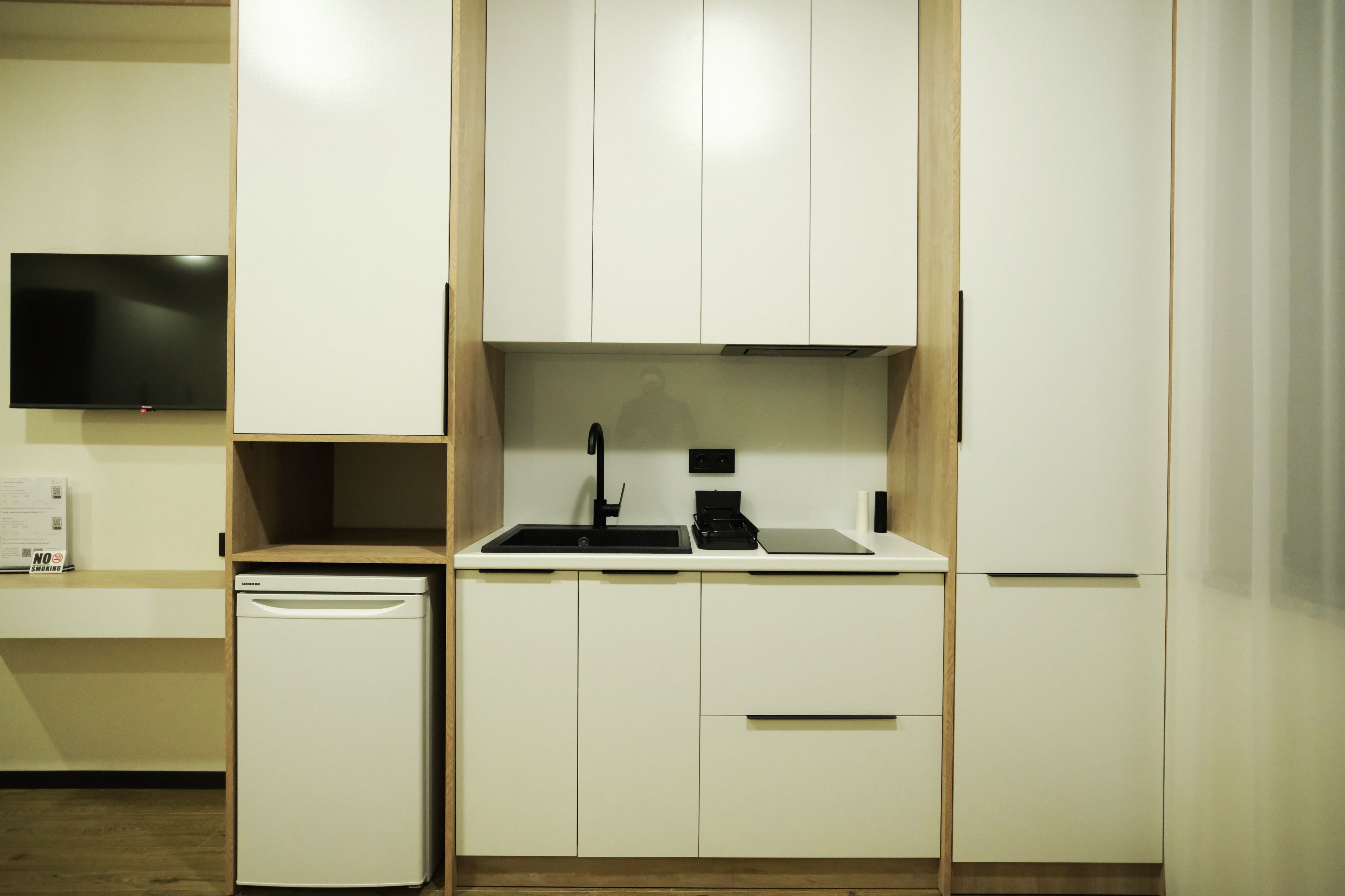 A compact kitchenette with a built-in dishwasher, a sink with a faucet, and a flat-screen TV mounted on the wall.